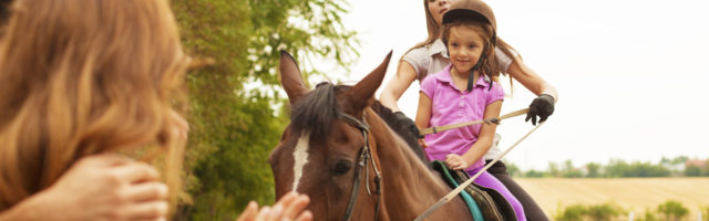 Child Riding Horse Outdoors. Little girl with riding hat learning how to ride a horse. Riding horse together with her instructor. Mother and father watching and encourage her. 