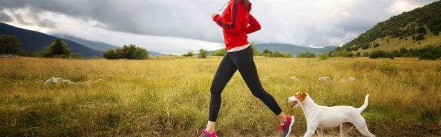 Jogging in the beautiful nature Young woman running in sports clothing with her pet dog. She is wearing a red sweater, doing exercise, in the beautiful nature, under the cloudy sky. Healthy lifestyle concept, keeping body in shape. Slightly toned image in analog / film colour. 